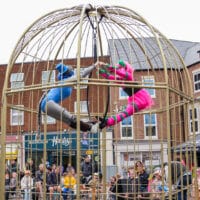 A wide shot of a large, dome-shaped metal cage set up in Gainsborough Market Place, with two street performers suspended inside. One performer in a bright pink costume with green accents and theatrical face paint hangs on the right, while another performer in a blue bodysuit with matching headpiece hangs on the left. Both hold onto a central hanging hoop, leaning outward with legs extended toward each other to form a balanced pose. Below and around the cage, a seated and standing audience watches, including families and children. The background shows brick buildings, shopfronts, and a market-style black canopy, placing the scene in a busy public street setting.