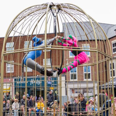A wide shot of a large, dome-shaped metal cage set up in Gainsborough Market Place, with two street performers suspended inside. One performer in a bright pink costume with green accents and theatrical face paint hangs on the right, while another performer in a blue bodysuit with matching headpiece hangs on the left. Both hold onto a central hanging hoop, leaning outward with legs extended toward each other to form a balanced pose. Below and around the cage, a seated and standing audience watches, including families and children. The background shows brick buildings, shopfronts, and a market-style black canopy, placing the scene in a busy public street setting.